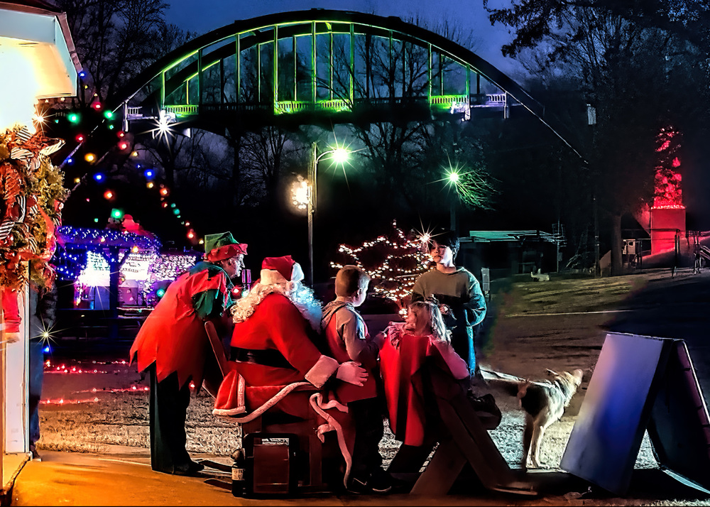 Santa, Mrs. Claus, elves sit in Santa's sleigh and talk to children with the Cotter Bridge in the background